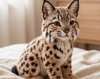 May include: A crocheted lynx figurine featuring tan and cream yarn with dark brown spots. The lynx has pointed ears with black tufts, large amber-colored eyes, a small pink nose, and white whiskers. It is depicted in a seated position.
