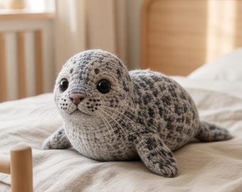 May include: A crochet seal plush toy featuring a speckled gray and white yarn pattern. The stuffed animal has large black plastic eyes, a small pink nose, and long thin whiskers extending from its snout, with a rounded body and flippers.