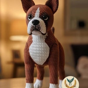 May include: A crocheted boxer dog figurine featuring brown yarn with a white chest, muzzle, and paws. The dog has dark, glossy eyes and a black nose. A circular logo with the text Promtiva Studio is visible in the lower right corner of the image.