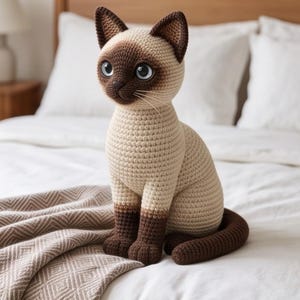 May include: A crocheted Siamese cat plush toy featuring a cream-colored body with dark brown ears, face mask, paws, and tail. The cat has large, round, light blue eyes, a small dark nose, and thin white whiskers, sitting in an upright position.