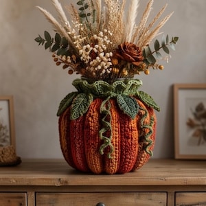 May include: A crochet pumpkin cozy in shades of orange and red, featuring green crocheted leaves and curly vines around the top rim. The cozy encases a glass vase filled with a dried floral arrangement of pampas grass, eucalyptus, and a brown rose.