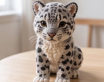 May include: A crocheted stuffed animal shaped like a snow leopard. The figure features a light gray body with dark gray and black spots, a cream-colored chest, and large brown eyes. It has white whiskers and small rounded ears with dark edges.
