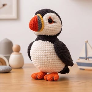 Può includere: Un peluche a forma di pulcinella di mare realizzato all'uncinetto con corpo nero e bianco, faccia bianca e un becco multicolore nelle tonalità del rosso, giallo e grigio. L'uccello ha zampe palmate arancioni e occhi scuri, con punti visibili.