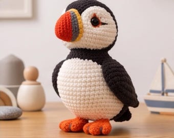 May include: A crochet puffin plush toy featuring a black and white body, a white face, and a multi-colored beak in shades of red, yellow, and grey. The bird has bright orange webbed feet and dark, round eyes, crafted with visible yarn stitches.