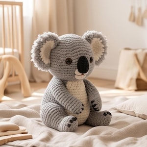 May include: A handmade crochet koala stuffed animal featuring a grey body with a cream-colored belly and inner ears. The koala has dark brown safety eyes, a black stitched nose, and small black claw details on its hands and feet, crafted in yarn.