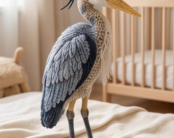 May include: A handcrafted crochet heron figurine featuring a textured plumage in shades of slate blue, grey, and cream. The bird has a long yellow beak, dark grey legs, and a crest of dark blue yarn fibers extending from the back of its head.