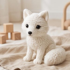 Könnte beinhalten: Ein weißes, gehäkeltes Fuchskuscheltier mit strukturiertem Garnkörper, spitzen Ohren und einem flauschigen Schwanz. Der Fuchs hat große, runde, dunkelbraune Augen, eine kleine schwarze Nase und dünne weiße Schnurrhaare und sitzt aufrecht.