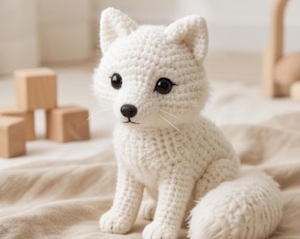 Puede incluir: Un zorro de peluche tejido a ganchillo en color blanco con cuerpo de hilo texturizado, orejas puntiagudas y una cola esponjosa. El zorro tiene ojos grandes, redondos y de color marrón oscuro, una nariz negra pequeña y bigotes blancos finos.