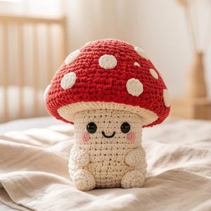 May include: A crocheted mushroom plush toy featuring a bright red cap with white spots and a cream-colored stem. The stem has a stitched smiling face with black eyes and pink cheeks. The mushroom has small arms and legs in a seated position.