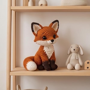 K&ouml;nnte beinhalten: Ein geh&auml;keltes Fuchskuscheltier aus orangefarbenem, wei&szlig;em und dunkelbraunem Garn. Der Fuchs hat gro&szlig;e schwarze Augen, spitze Ohren mit dunklen Spitzen und einen buschigen Schwanz mit wei&szlig;er Spitze. Er sitzt neben einem kleinen, beigen H&auml;kelhasen.
