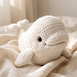 May include: A cream-colored, crocheted beluga whale plush toy featuring a rounded head, a small tail, two side fins, and two small, round, black plastic eyes. The texture of the yarn is visible throughout the entire body of the stuffed animal.