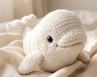 May include: A cream-colored, crocheted beluga whale plush toy featuring a rounded head, a small tail, two side fins, and two small, round, black plastic eyes. The texture of the yarn is visible throughout the entire body of the stuffed animal.