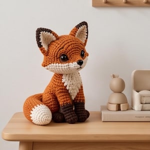 Könnte beinhalten: Ein gehäkeltes Fuchskuscheltier mit leuchtend orangefarbenem Körper, weißer Brust und weißer Schwanzspitze sowie dunkelbraunen Ohren und Pfoten. Der Fuchs hat große, glänzende schwarze Augen und eine kleine schwarze Nase und sitzt mit eingerolltem Schwanz.