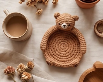 May include: A light brown crochet bear-shaped tray with a circular base, featuring a bear head at the top, two small arms on the sides, and two feet at the bottom. The tray is constructed from thick yarn in a spiral pattern with a white muzzle.