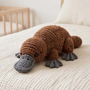 Puede incluir: Un ornitorrinco de peluche tejido a ganchillo en color marrón, con cuerpo de hilo texturizado, ojos redondos oscuros y un pico plano tejido en gris. La criatura tiene cuatro patas palmeadas grises y una cola ancha y plana con puntos visibles.