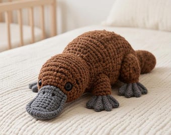May include: A brown crocheted platypus stuffed animal featuring a textured yarn body, dark round eyes, and a flat, grey crocheted bill. The creature has four grey webbed feet and a wide, flat tail, all crafted with visible yarn stitches.