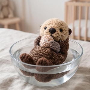 Pode incluir: Um brinquedo de lontra em crochê com cabeça bege e corpo castanho escuro, apresentando olhos pretos e nariz escuro. A lontra segura uma pequena bola redonda e texturizada bege e branca enquanto está sentada numa taça de vidro com água.