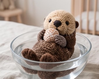 May include: A crocheted otter toy with a tan head and dark brown body, featuring black eyes and a dark nose. The otter is holding a small, round, textured beige and white ball while sitting inside a clear glass bowl filled with water.
