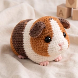 May include: A crocheted guinea pig plush toy featuring a tri-color pattern of brown, white, and dark brown yarn. The stuffed animal has black plastic safety eyes, a small pink stitched nose, and tiny pink crocheted feet, crafted with a dense stitch.