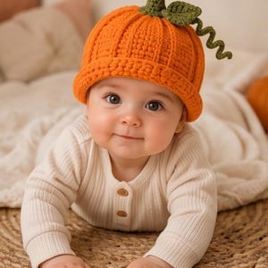Puede incluir: Un gorro de ganchillo de color naranja brillante con forma de calabaza, que presenta una textura acanalada, un borde enrollado y un tallo verde con una hoja y un zarcillo rizado en la parte superior. Se lleva con una camisa de canalé color crema.