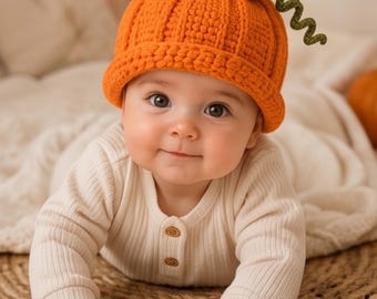 May include: A bright orange crochet hat shaped like a pumpkin, featuring a ribbed texture, a rolled brim, and a green stem with a leaf and a curly vine on top. The hat is worn with a long-sleeved, cream-colored ribbed shirt with two small buttons.