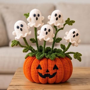 May include: A crocheted orange pumpkin with a black jack-o'-lantern face, serving as a base for five small, white, crocheted ghost figures attached to green stems with leaves, creating a decorative arrangement of yarn-based Halloween decor.