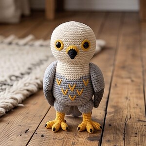 May include: A crochet bird toy featuring a cream-colored head, a gray body with yellow V-shaped embroidery, dark eyes with yellow rings, a black beak, and bright yellow feet with black-tipped claws. The stuffed animal stands upright on its own.