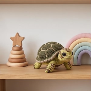 Könnte beinhalten: Eine gehäkelte Schildkröte mit einem dunkel olivgrünen Panzer mit hellbeigen Nähten, einem hellgrünen Kopf und Gliedmaßen mit braunen Flecken sowie großen schwarzen Augen. Die Schildkröte steht auf einem Holzregal neben einem hölzernen Stapelspielzeug.