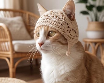 Könnte beinhalten: Eine cremefarbene Häkelhaube für eine Katze mit einem komplizierten Spitzenmuster, Ohröffnungen und kleinen baumelnden Bommeln an den Seiten. Die Haube wird von einer orange-weißen getigerten Katze mit hellen Augen getragen.