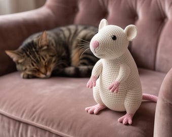 May include: A cream-colored crocheted rat toy with a pink nose, pink hands, and pink feet. The stuffed animal has small black eyes, rounded ears, and a long tail, standing upright with a textured yarn surface and visible stitching throughout.