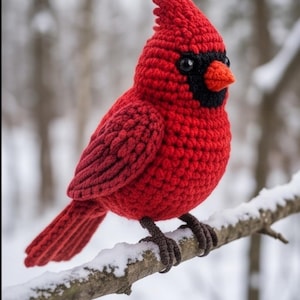 Op de afbeelding: Een gehaakt kardinaalvogel-figuurtje gemaakt van felrood garen, met een zwart masker rond de ogen, een kleine oranje snavel en donkerbruine garenpootjes. De vogel heeft een getextureerd, geknoopt uiterlijk en zit op een dunne, met sneeuw bedekte tak.