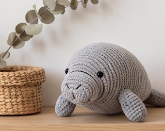 May include: A gray crocheted manatee plush toy featuring a rounded body, two side flippers, a flat tail, and small black bead eyes. The surface of the toy is crafted with a consistent yarn stitch pattern, and it has a stitched mouth detail.