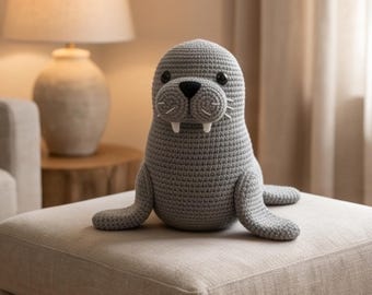 May include: A gray crocheted walrus plush toy featuring a rounded body, two white tusks, a black stitched nose, and dark circular eyes. The surface of the toy displays a consistent yarn texture with visible stitches throughout the entire form.