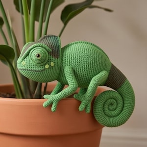 May include: A handcrafted crochet chameleon toy featuring a vibrant green body with dark grey accents on the head and tail. The stuffed animal has a textured, stitched surface, a large circular eye, and a curled tail resting on the edge of a pot.