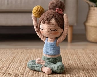 Puede incluir: Una muñeca de ganchillo con el pelo castaño recogido en un moño, que lleva un top corto azul claro y mallas verde salvia, sentada en posición de loto con los brazos levantados sobre la cabeza sosteniendo una pequeña pelota amarilla.