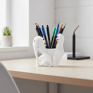 Puede incluir: Un organizador de escritorio blanco con forma de cráneo de dinosaurio lleno de bolígrafos y lápices. El accesorio de escritorio está hecho de un material duro y liso y tiene un diseño detallado. Los bolígrafos y lápices son de varios colores.