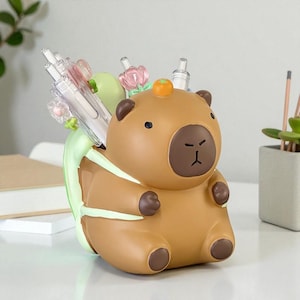 Puede incluir: Un organizador de escritorio con forma de carpincho marrón con una parte trasera verde claro y una pequeña fruta naranja en la cabeza. El organizador contiene bolígrafos y lápices, con algunas flores decorativas. El carpincho tiene una cara y patas de color marrón oscuro.