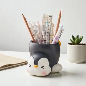 Puede incluir: Un organizador de escritorio con forma de pingüino en negro, blanco y amarillo, que contiene lápices, bolígrafos y una regla. El recipiente tiene una cara sonriente y mejillas rosadas. Una pequeña planta suculenta en una maceta blanca está en el fondo.