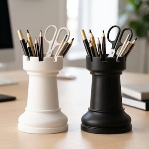 Op de afbeelding: Twee bureaulade-organizers in de vorm van schaaktorens, één witte en één zwarte, elk gevuld met potloden, pennen en scharen. De witte toren bevat een witte schaar, terwijl de zwarte toren een zwarte schaar bevat. De bureau-accessoires staan op een licht houten oppervlak.