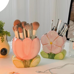 Puede incluir: Dos porta-brochas de maquillaje de cerámica con forma de flores. Uno tiene forma de tulipán, el otro de flor. Ambos son rosas con detalles verdes y amarillos. Los soportes están llenos de brochas de maquillaje.