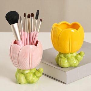 Op de afbeelding: Twee keramische borstelhouders in de vorm van een tulp. Eén is roze en bevat make-up borstels. De andere is geel. Beide staan op groene, getextureerde bases. De houders staan op een wit oppervlak.