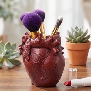 Puede incluir: Un soporte para brochas de maquillaje en forma de corazón anatómico de color burdeos. El soporte contiene varios pinceles de maquillaje con cerdas moradas y blancas. Una pequeña maceta de terracota con una planta suculenta está en el fondo.