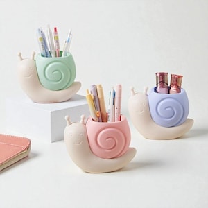 May include: Three pastel-coloured snail-shaped desk tidies. Each snail features a beige body and a spiral shell in green, pink, or blue. The organisers hold pens, pencils, and makeup items, adding a playful touch to any workspace.