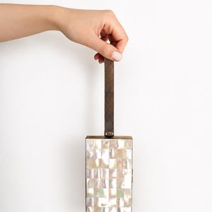Può includere: Una pochette rettangolare caratterizzata da un mosaico di tessere in madreperla bianca iridescente. La borsa presenta una cornice in metallo dorato e un sottile cinturino da polso in maglia metallica scura fissato al centro superiore.