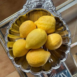 Könnte beinhalten: Eine silberfarbene, verzierte Servierschale gefüllt mit sechs leuchtend gelben Zitronen. Die Schale hat einen gewellten Rand und einen goldfarbenen Innenraum. Die Zitronen sind reif und haben eine strukturierte Oberfläche.