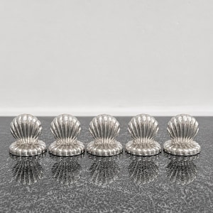 Può includere: Un set di cinque poggiacoltelli in metallo argentato a forma di conchiglia. Ogni pezzo presenta un design a ventaglio smerlato con creste verticali che si irradiano dalla base, creando un aspetto uniforme per tutti e cinque gli oggetti.