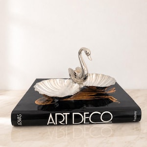 Pode incluir: Um prato de metal prateado em forma de cisne com duas taças recortadas em forma de concha que se estendem da figura central. O objeto repousa sobre um livro preto com o texto ART DECO impresso em branco na lombada e na capa frontal.