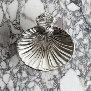 Può includere: Un piatto argentato a forma di conchiglia con un manico decorativo. Il piatto ha un design a coste che si irradia dal centro. Il manico presenta un motivo a vortice. Il piatto è esposto su una superficie di marmo.