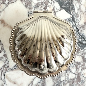 Puede incluir: Una caja de baratijas plateada en forma de concha con tapa con bisagras. La caja tiene un diseño detallado y acanalado y está bordeada con pequeñas cuentas decorativas. La caja está cerrada y refleja el entorno.