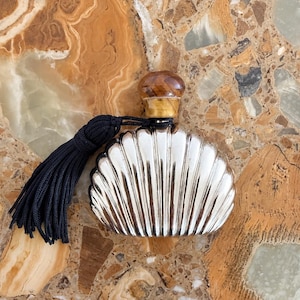 Peut inclure: Un flacon de parfum argenté en forme de coquillage avec des stries verticales. Le flacon est doté d'un bouchon rond en pierre polie brune et d'un long pompon en tissu bleu marine foncé attaché au col par un cordon fin.